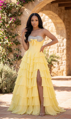 A woman stands outdoors before a stone archway and lush greenery, wearing the A-Line Ruffle Prom Gown: PromGirl Exclusive 250203—a strapless long prom dress with a beaded sweetheart neckline and high slit.