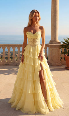 A woman wears the Long Ruffle Prom Gown: PromGirl Exclusive 250205 in light yellow, featuring a strapless sweetheart bodice and ruffled skirt, as she stands on a stone terrace overlooking water and greenery beneath a clear sky.