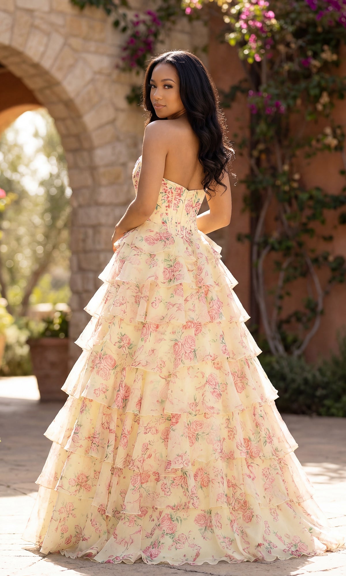A woman stands outdoors in an A-Line Print Prom Gown: PromGirl Exclusive 250206, gazing over her shoulder. A stone archway and lush greenery provide a dreamy setting for this elegant floral-print dress.