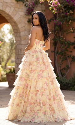 A woman stands outdoors in an A-Line Print Prom Gown: PromGirl Exclusive 250206, gazing over her shoulder. A stone archway and lush greenery provide a dreamy setting for this elegant floral-print dress.