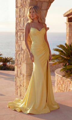 A woman wearing the Pleated Long Prom Dress: PromGirl Exclusive 250219 in light yellow stands outdoors by a stone archway, with palm plants and the sea in the background. The dress features thin straps and a thigh-high slit.