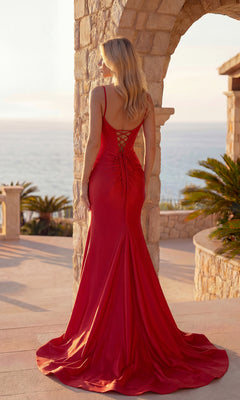 A woman in the Pleated Long Prom Dress: PromGirl Exclusive 250219 stands outdoors by a stone archway, facing away with the ocean behind her. She wears a red satin gown with a lace-up back.