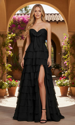 A woman wears the black PromGirl Exclusive Long Prom Dress 250233B—strapless, with a tiered ruffle skirt, front bow, and thigh-high slit—as she stands in a sunlit courtyard surrounded by potted plants and flowers.