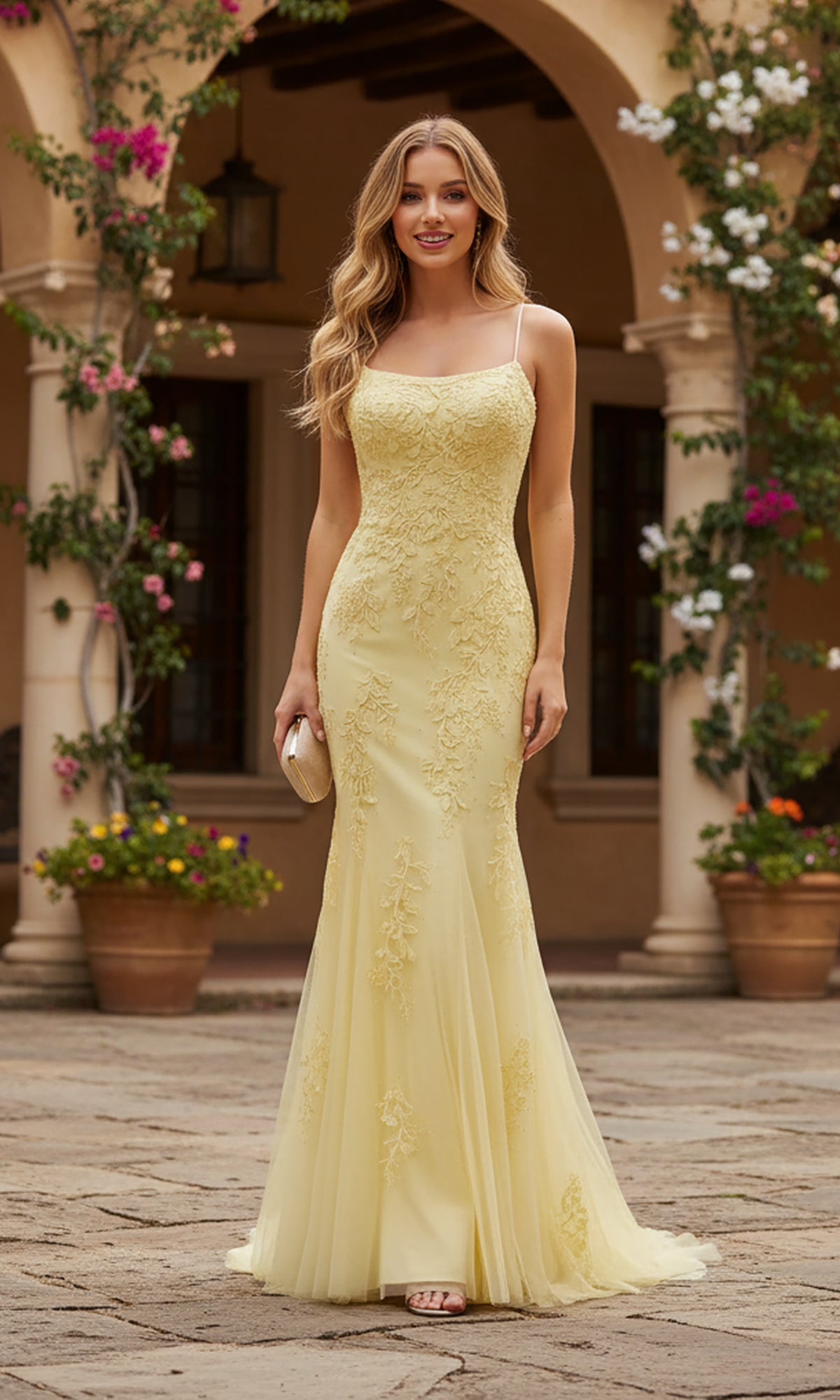 Wearing the PromGirl Exclusive 250226, a pale yellow, floor-length lace prom dress with a train, a woman stands outdoors on stone pavement holding a clutch, surrounded by flowering plants and an arched building.