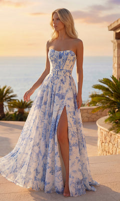 A woman in the PromGirl Exclusive 250236 Floral Chiffon Prom Dress in Ivory/Blue Print stands outdoors by palm plants, with an ocean sunset providing a stunning backdrop.