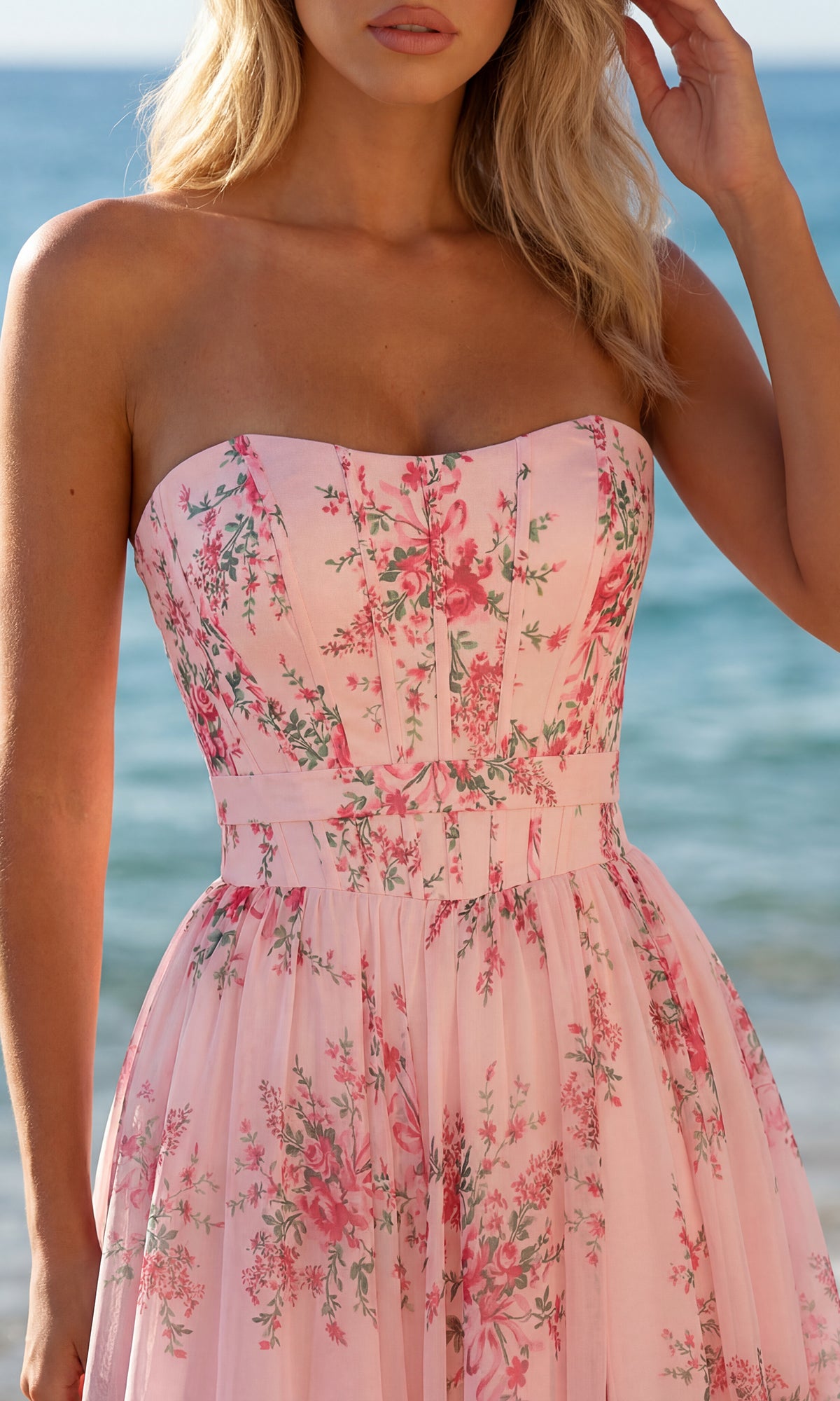 Woman wearing the Floral Chiffon Prom Dress: PromGirl Exclusive 250236, featuring a pink floral print, stands by the ocean with waves in the background.