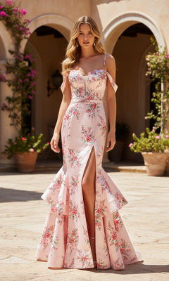 A woman in the PromGirl Exclusive 250237 long prom dress, featuring a pink print satin fabric with a sweetheart neckline, mermaid skirt, and front slit, stands outdoors near potted plants and arched doorways.