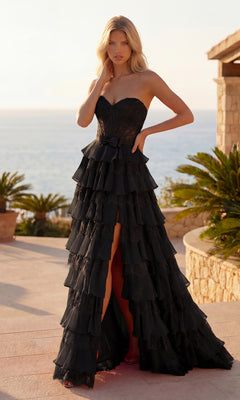 A woman in the black Chiffon and Lace Prom Gown: PromGirl Exclusive 250238 stands outdoors by stone architecture, with palm plants and the ocean visible in the background.