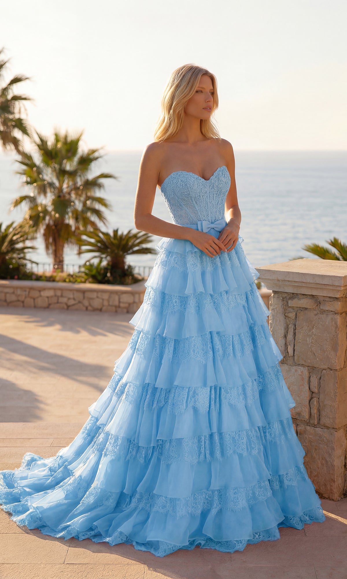 A woman in a light blue Chiffon and Lace Prom Gown (PromGirl Exclusive 250238) stands outdoors by a stone wall, with palm trees and the ocean in the background.