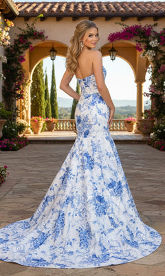 A woman stands on a stone patio with arches and flowers, wearing the PromGirl Exclusive 250239—a strapless blue and white floral mermaid prom dress featuring a corset bodice.