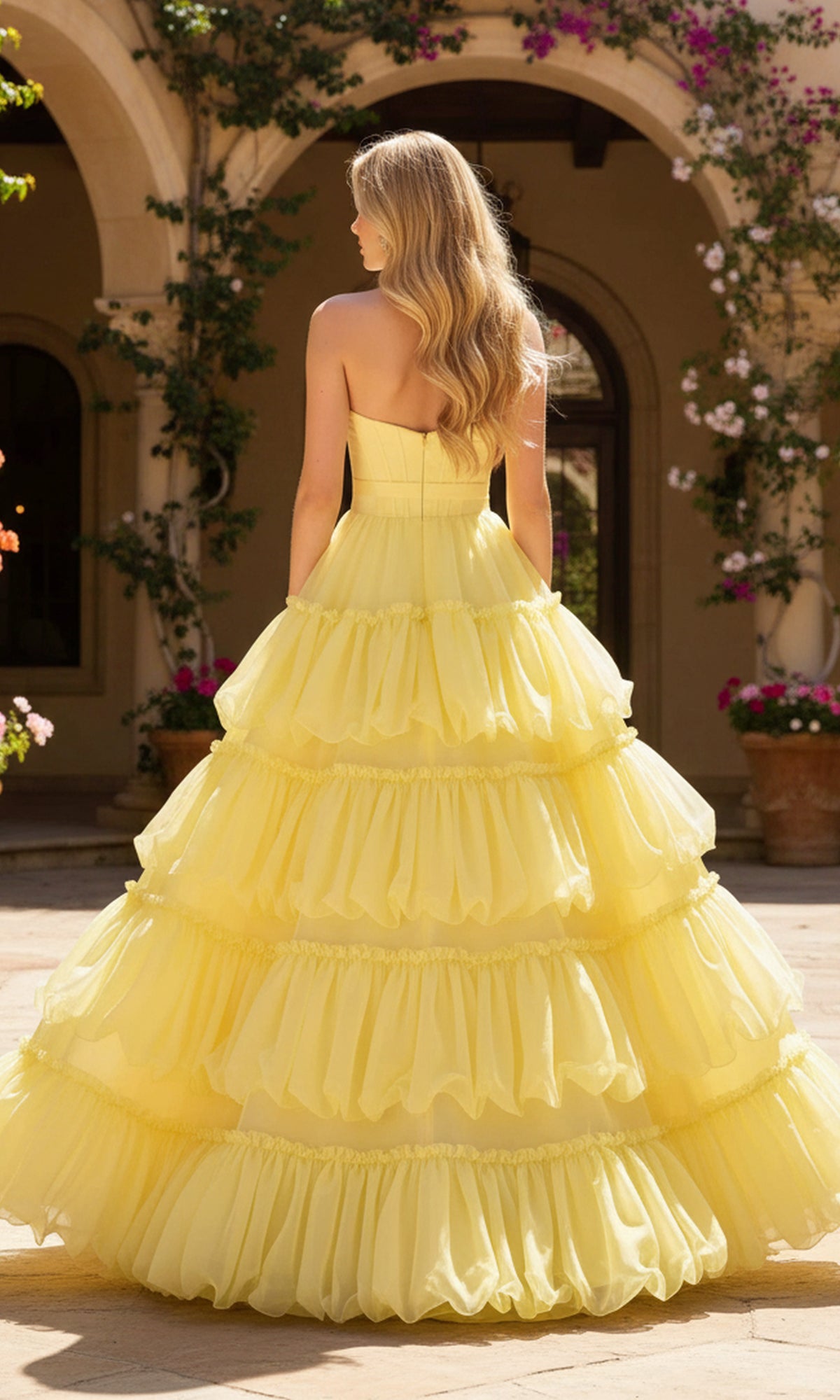 A woman with long blonde hair wears the PromGirl Exclusive 250240B long prom dress while standing outside in a courtyard surrounded by flowering plants.