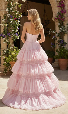 A woman stands outdoors by arches and flowers, modeling the PromGirl Exclusive 250240B—a pink, strapless long prom dress with graceful tiered layers—as she poses with her back to the camera.
