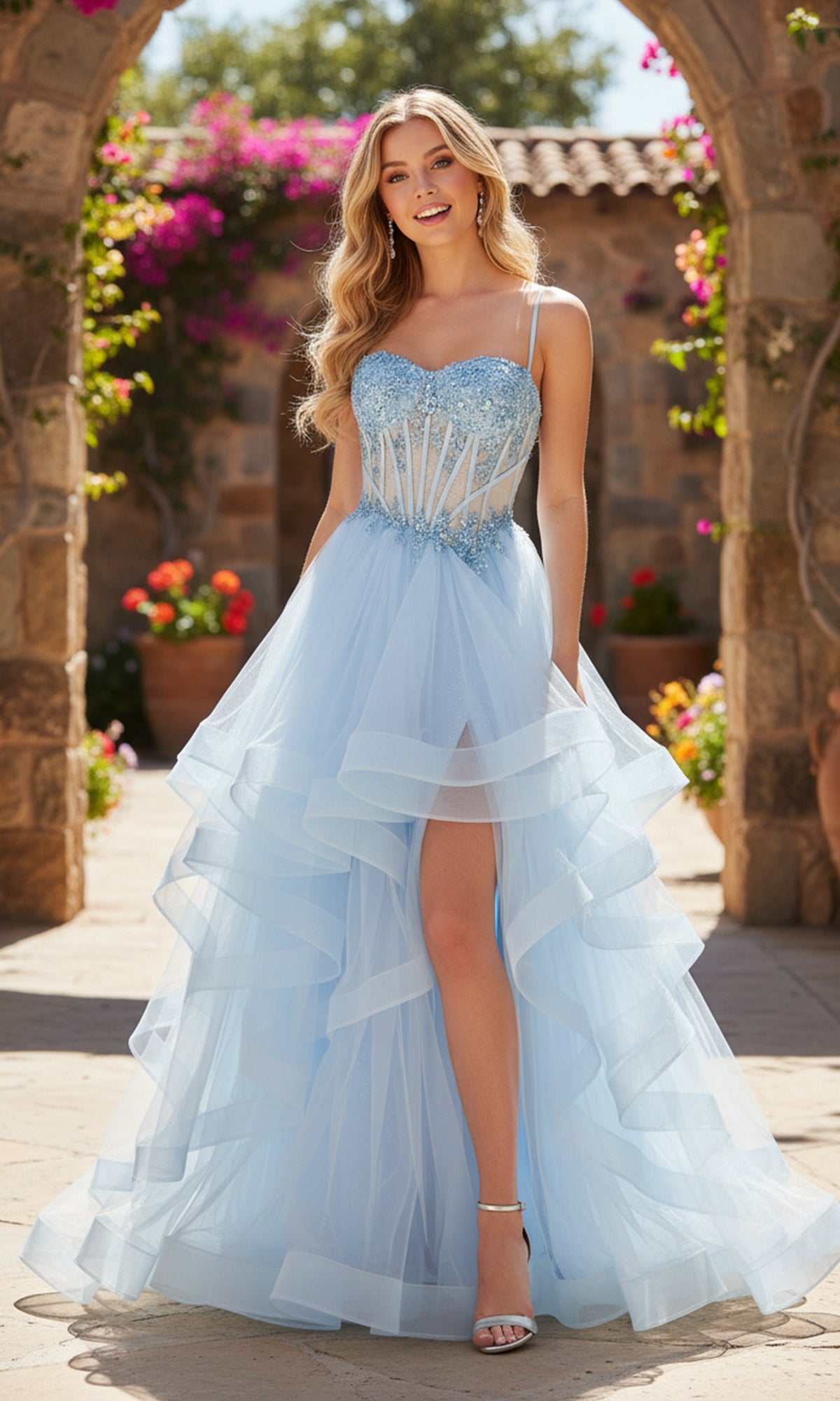 A woman in a light blue Long Prom Dress: PromGirl Exclusive 250241 with beaded corset, layered tulle skirt, and high slit stands outdoors in a sunlit garden framed by stone arches and blooming flowers.