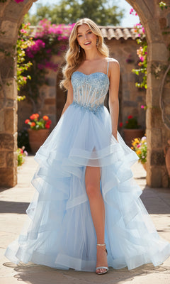 A woman in a light blue Long Prom Dress: PromGirl Exclusive 250241 with beaded corset, layered tulle skirt, and high slit stands outdoors in a sunlit garden framed by stone arches and blooming flowers.