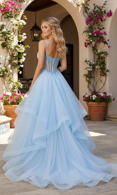 A woman wearing the Long Prom Dress: PromGirl Exclusive 250241, featuring a light blue layered tulle skirt and a beaded corset bodice, stands outdoors by potted flowers and a white wall.