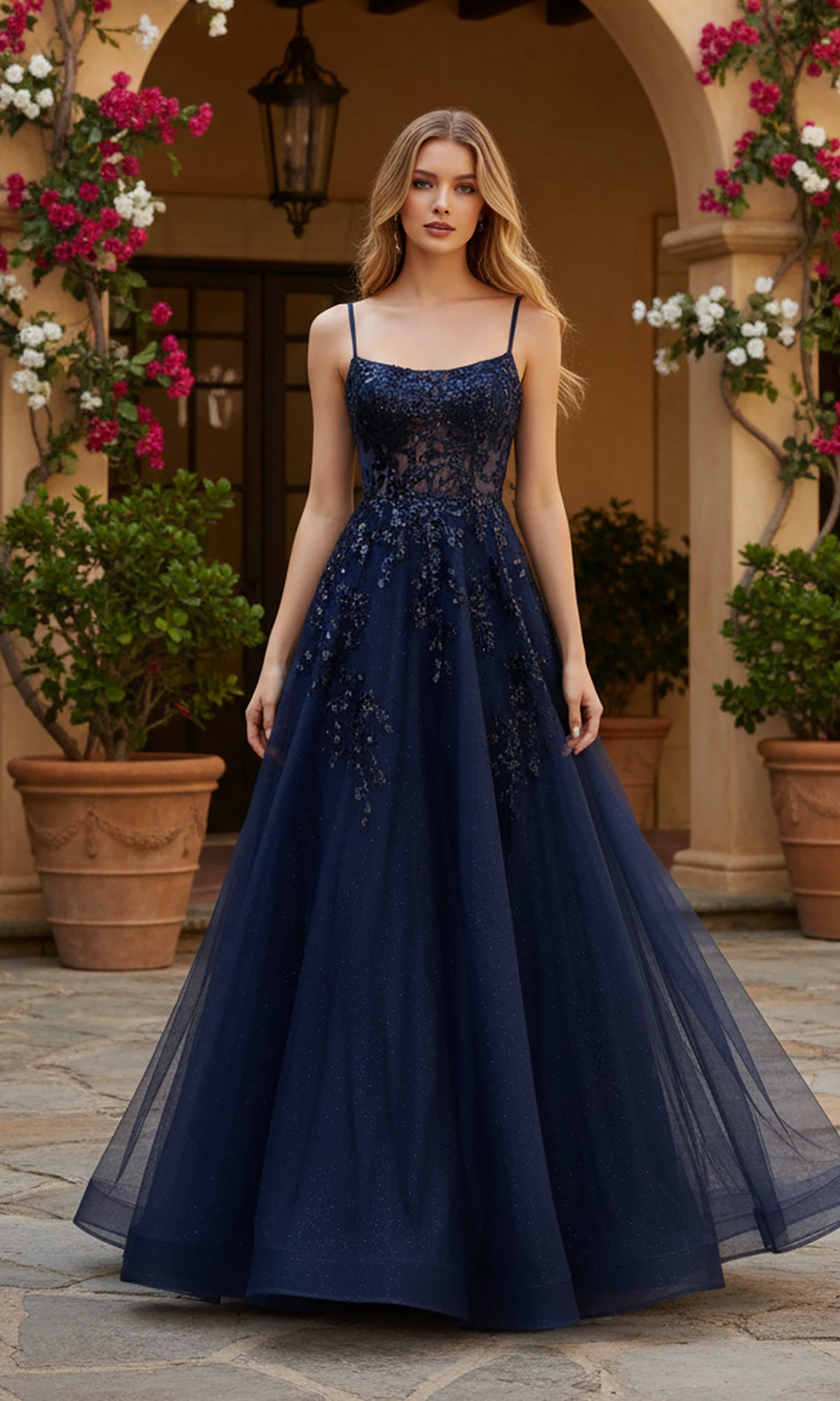 A woman stands outdoors by potted plants, wearing the PromGirl Exclusive 250245 Long Prom Dress—a floor-length navy gown with thin straps, floral bodice detailing, and an adjustable lace-up back.