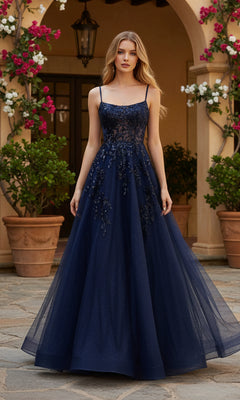 A woman stands outdoors by potted plants, wearing the PromGirl Exclusive 250245 Long Prom Dress—a floor-length navy gown with thin straps, floral bodice detailing, and an adjustable lace-up back.