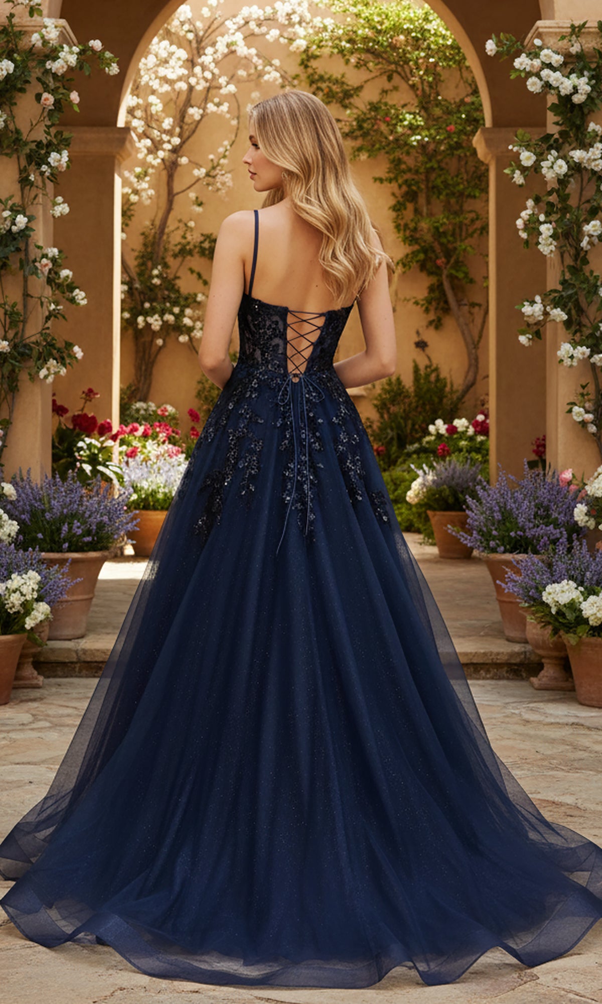 A woman wearing the PromGirl Exclusive 250245, a navy blue, floor-length gown with an adjustable lace-up back, stands in a garden courtyard surrounded by potted flowering plants.