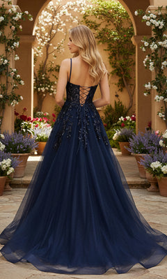 A woman wearing the PromGirl Exclusive 250245, a navy blue, floor-length gown with an adjustable lace-up back, stands in a garden courtyard surrounded by potted flowering plants.