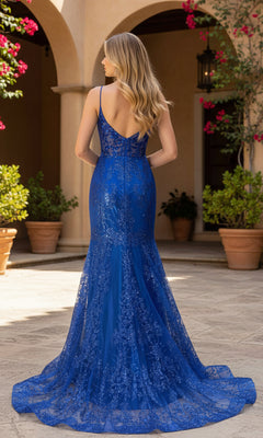 A woman wearing the PromGirl Exclusive 250249 long prom dress in blue lace with thin straps stands outside among potted plants and Mediterranean-style architecture, facing away.
