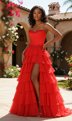 A woman wearing the PromGirl Exclusive 250267, a strapless ruffled long prom dress with a thigh-high slit, stands outdoors on a sunny patio, surrounded by flowers and Mediterranean-style architecture.