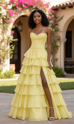 Wearing the PromGirl Exclusive 250267, a strapless yellow ruffled long prom dress with a tiered skirt and thigh-high slit, a woman stands outdoors before arched doorways and blooming flowers, embodying PromGirl elegance.