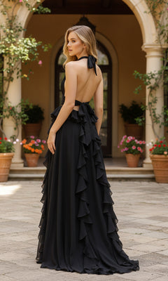 A woman wearing the PromGirl Exclusive 250269—a floor-length black chiffon prom dress with a halter neckline and open back—stands outdoors in front of an arched doorway and potted plants.