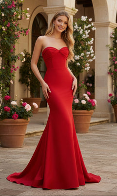 A woman in a red strapless mermaid Long Prom Dress: PromGirl Exclusive 250270 with back zipper closure stands outdoors on a stone patio, surrounded by potted flowers and an arched building in the background.
