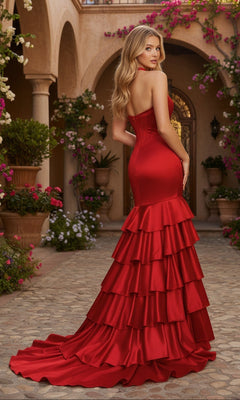 Wearing the PromGirl Exclusive 250294, a woman stands outdoors in a strapless, form-fitting red mermaid gown with tiered ruffles, surrounded by flowering plants and arched architecture.
