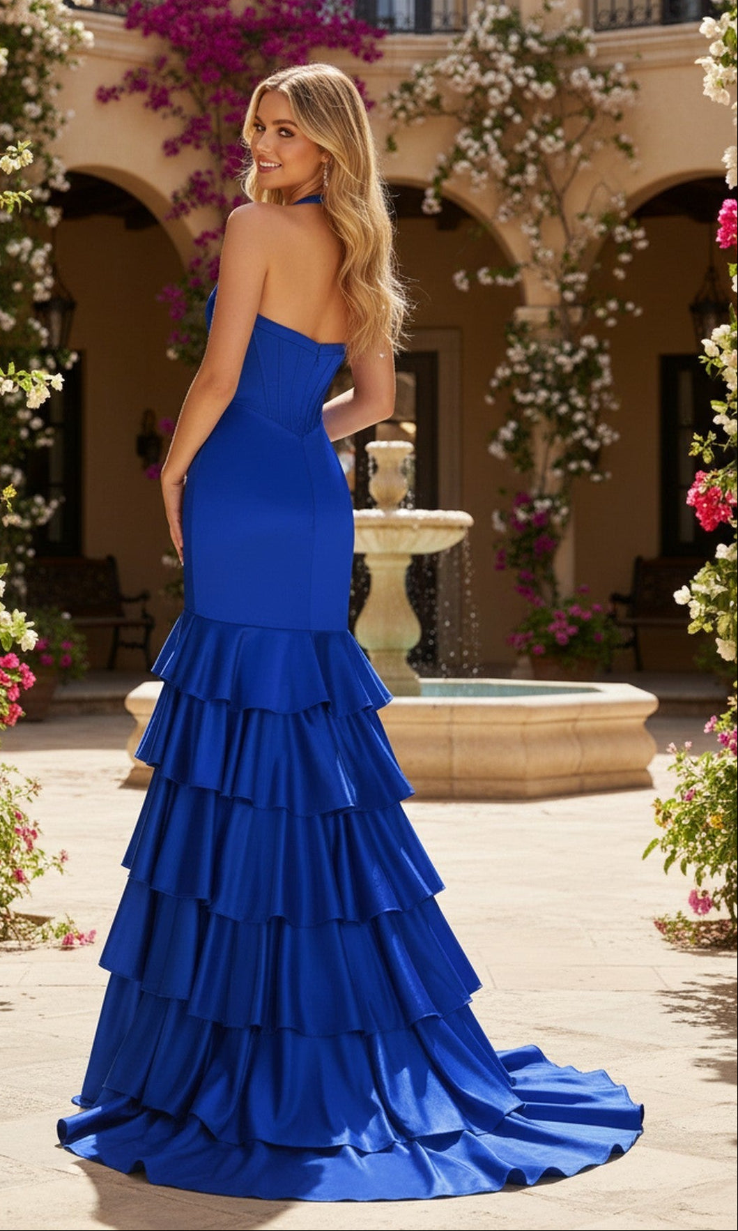 A woman wearing the Long Prom Dress: PromGirl Exclusive 250294, a strapless royal blue gown with a corset bodice and tiered ruffles, stands in a courtyard with a stone fountain and blooming flowers.