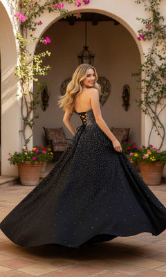 Wearing the PromGirl Exclusive 250297, a woman in a black strapless satin gown with an a-line skirt and white sparkles stands in an arched courtyard, turning slightly and smiling over her shoulder.
