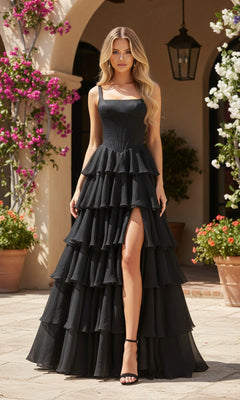 A woman in a black PromGirl Exclusive 250300 long prom dress with a corset bodice, tiered chiffon skirt, and thigh-high slit pairs it with black heels and stands outdoors near potted plants and flowering vines.