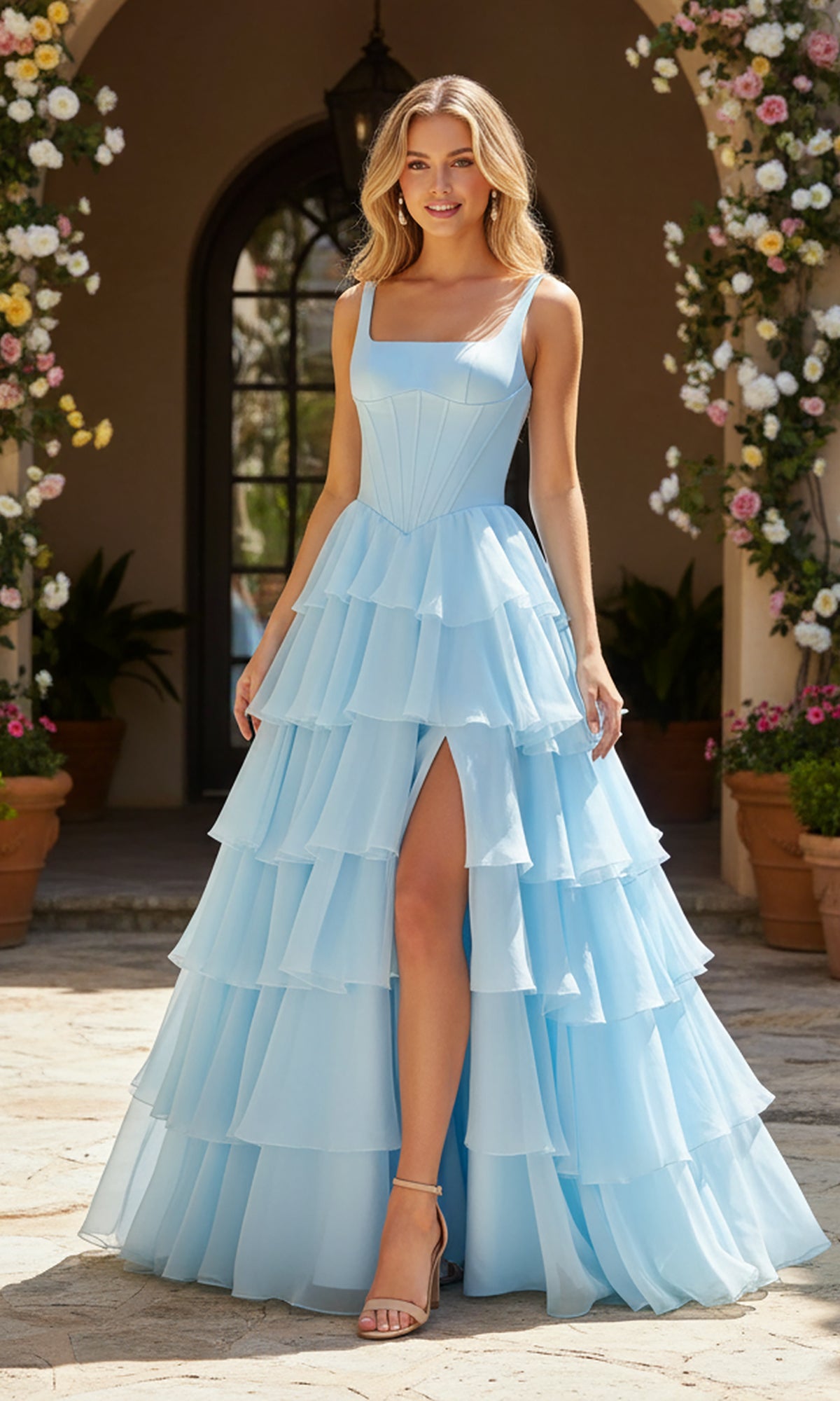 A woman wearing the PromGirl Exclusive 250300 long prom dress in light blue, featuring a corset bodice, tiered ruffle skirt, and square neckline, stands outdoors by potted flowers and an arched doorway.