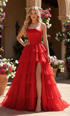 A woman stands outdoors in the PromGirl Exclusive 250300, a floor-length red prom dress with a tiered chiffon skirt and high slit, surrounded by potted flowers.