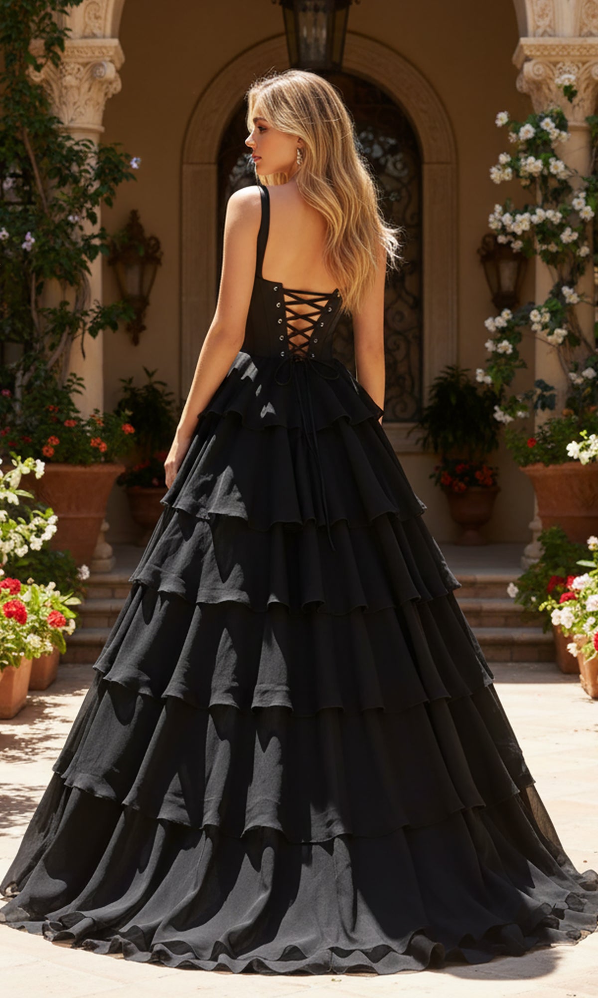 A woman wearing the PromGirl Exclusive 250300, a long black prom dress with a corset bodice and tiered chiffon skirt, stands outdoors by stone steps and potted plants.