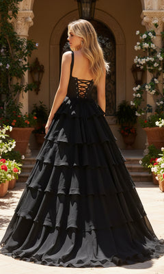 A woman wearing the PromGirl Exclusive 250300, a long black prom dress with a corset bodice and tiered chiffon skirt, stands outdoors by stone steps and potted plants.