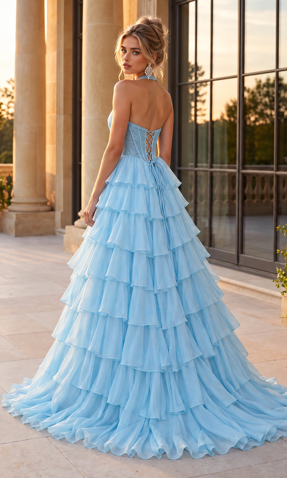 A woman stands on a stone terrace by large windows, looking over her shoulder, wearing the Halter Prom Ball Gown: PromGirl Exclusive 250352, featuring a delicate corset bodice and a tiered ruffle skirt.