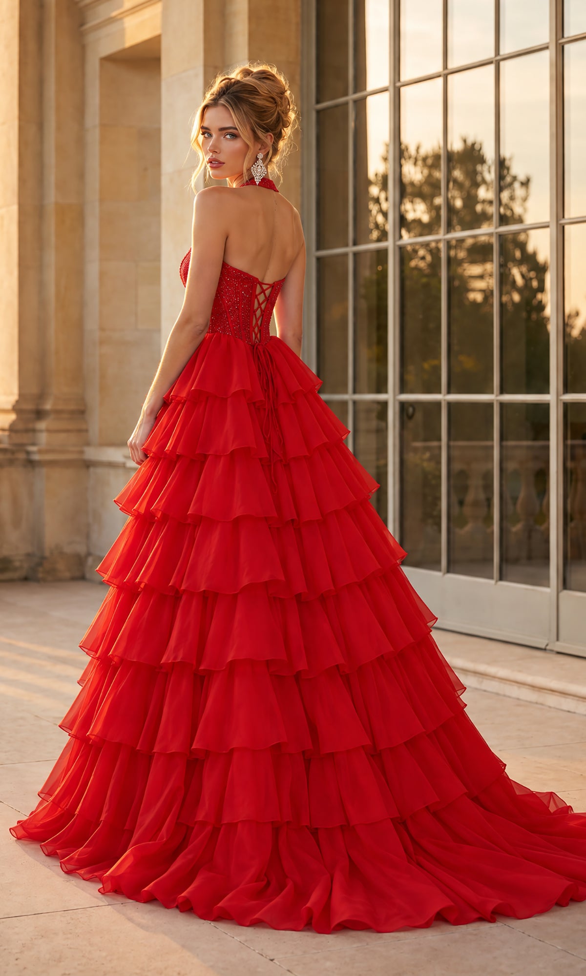 A woman in the Halter Prom Ball Gown: PromGirl Exclusive 250352 stands outdoors, looking over her shoulder, with large windows and stone columns in the background.
