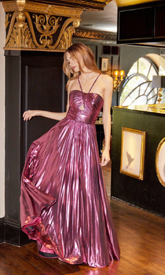 A woman indoors near a decorated column holds the side of her Long Metallic A-Line Prom Dress, Juliet JT2503A, featuring a shiny strappy-back design in metallic pink.