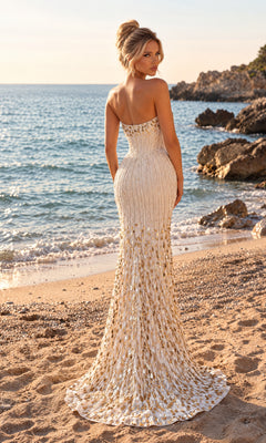 A woman stands on a sandy beach facing the ocean, wearing the PromGirl Exclusive 250545 long prom dress—a strapless gown with a fitted gold and white design—while rocks and waves form the background.