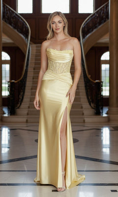 A woman in the Poly USA 25056 long yellow prom dress, featuring a strapless lace corset bodice and thigh-high slit, stands in a grand foyer with a staircase behind her.