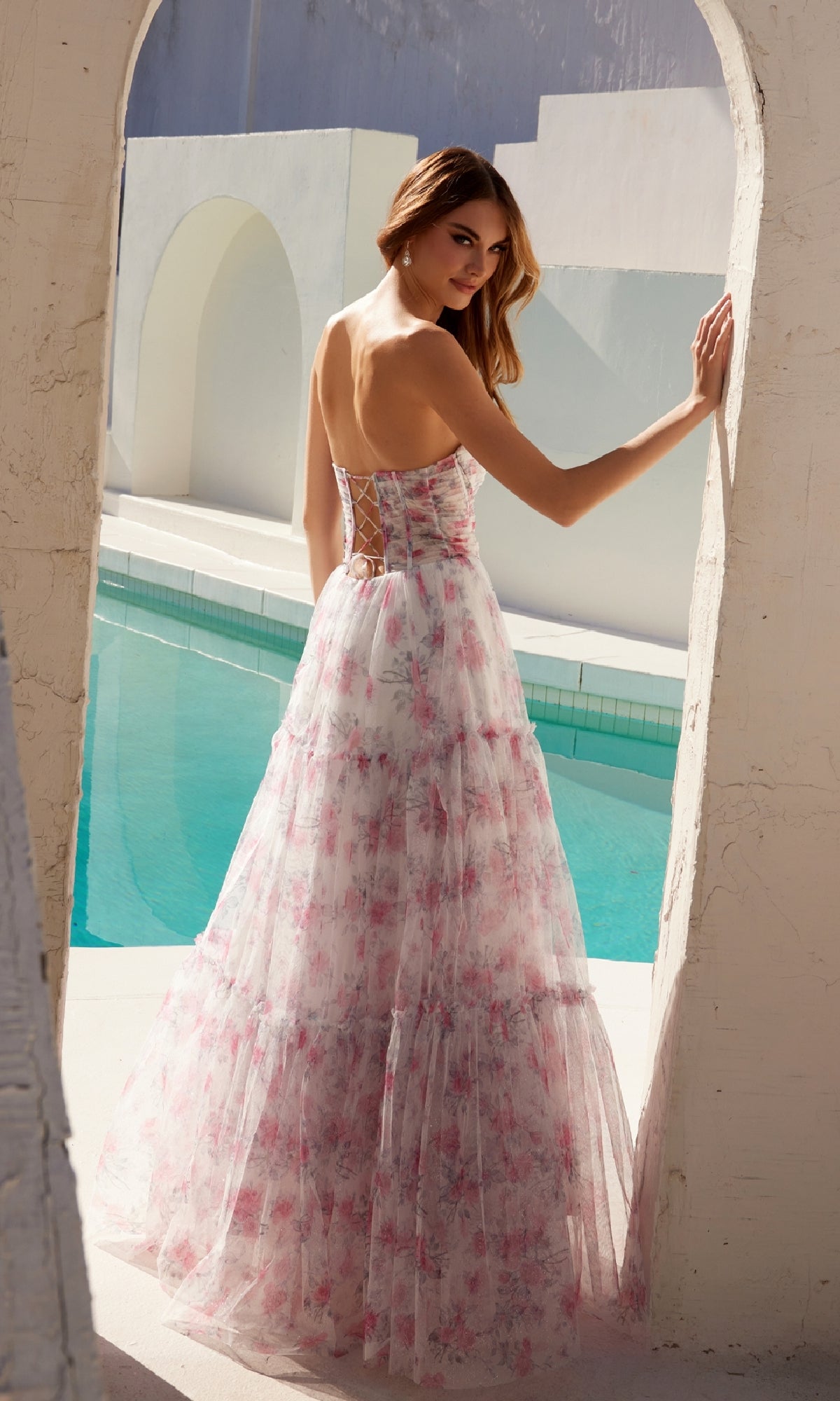 A woman wearing the Strapless Long Print Prom Dress Juliet JT2507A stands by a bright blue pool under an archway, glancing over her shoulder.