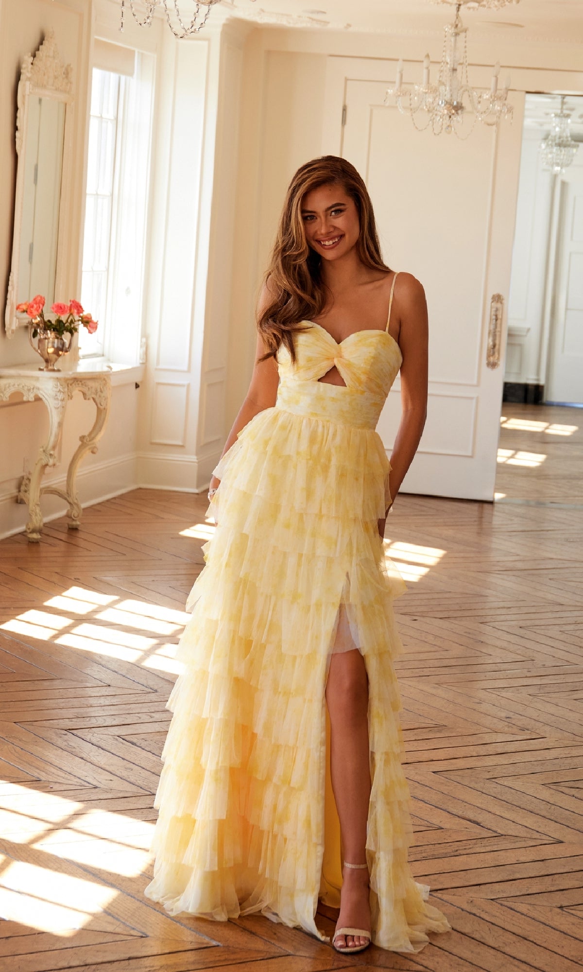 A woman smiles at the camera in a bright room, wearing the Long Yellow Ruffled Prom Dress: Juliet JT2508A, featuring spaghetti straps, a high slit, and a tiered A-line skirt.