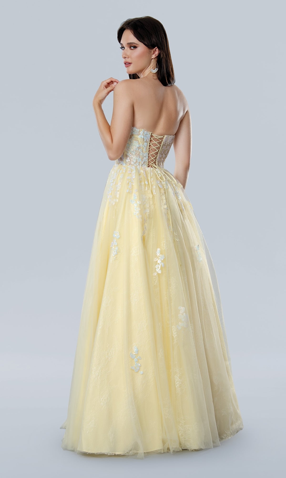 A woman models the Long Prom Dress: Stella Couture 25108, showcasing its strapless lace design with floral embroidery and a lace-up back as she stands facing away.