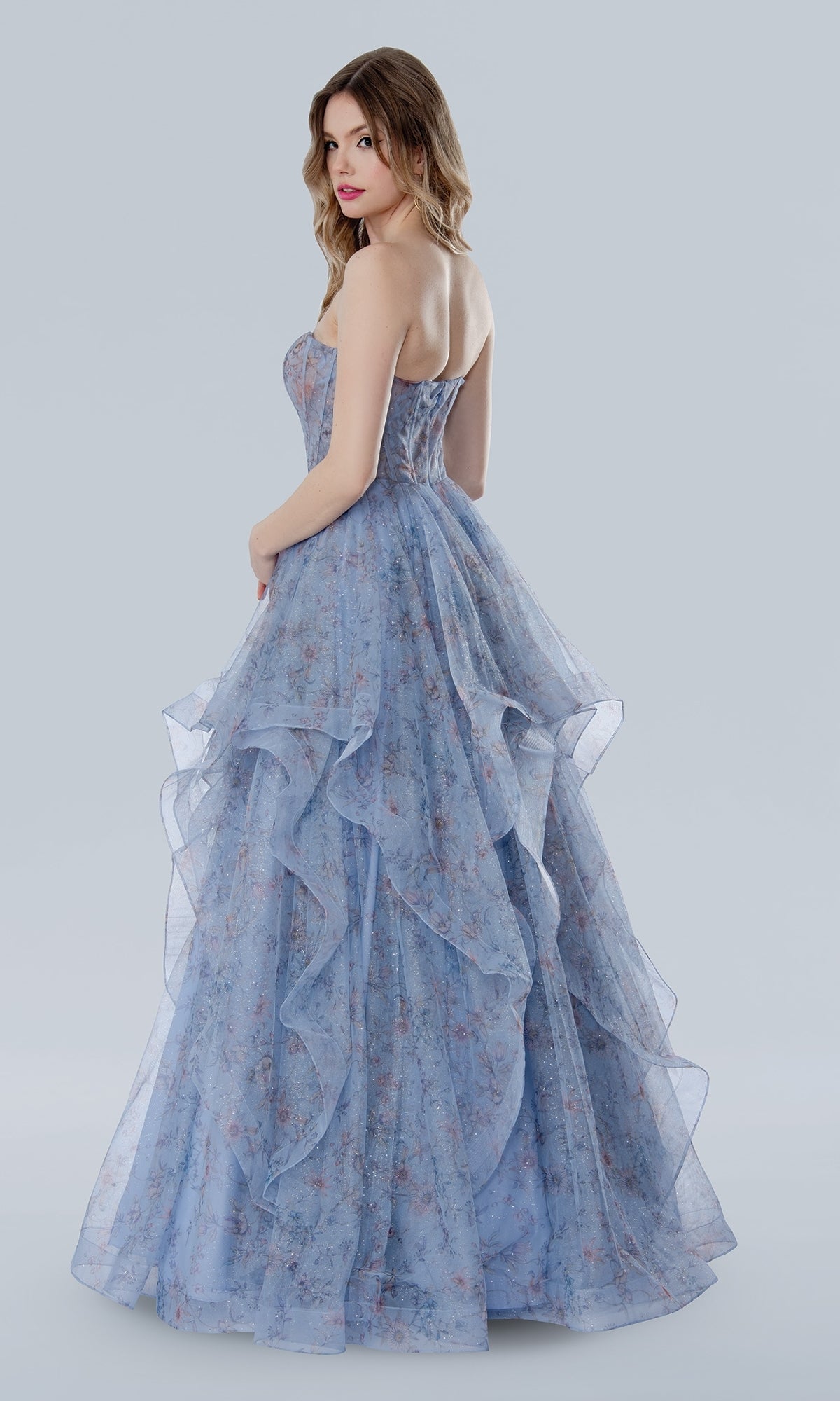 A woman models the Stella Couture 25119 floral prom ball gown, a strapless dress with a corset bodice, light blue floral pattern, and layered ruffles, set against a plain light gray background.