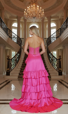 A woman in the Poly USA 25162 long prom dress with a bright pink, strapless, tiered A-line skirt stands at the base of a grand, chandelier-lit staircase.
