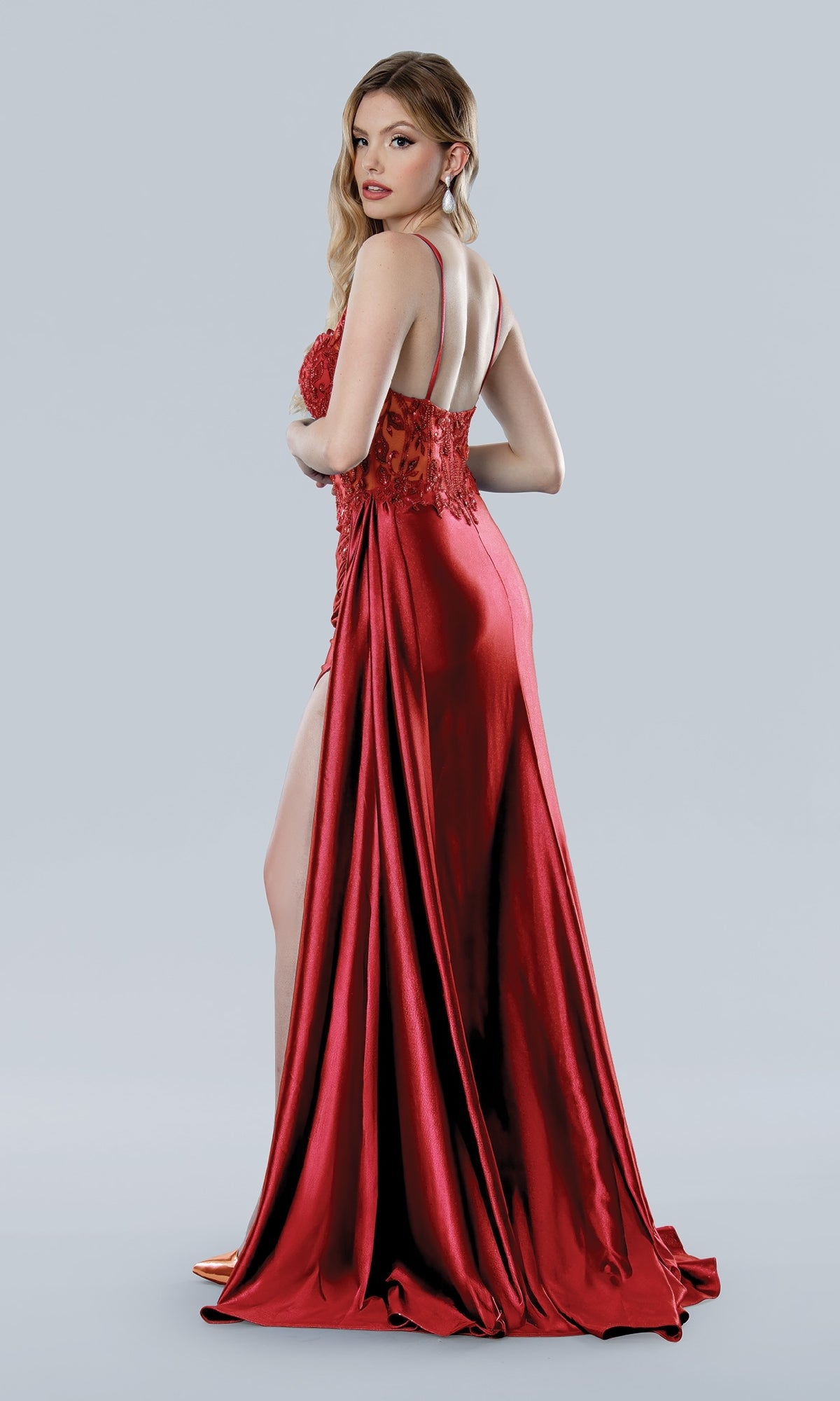 A woman models the Stella Couture 25166 long red prom dress with a high leg slit, standing sideways and looking over her shoulder against a plain light gray background.