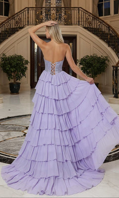 A woman stands with her back to the camera in an ornate foyer, holding the Long Prom Dress: Poly USA 25168—a strapless chiffon gown with a tiered ruffle skirt—on marble floors beside a curved staircase.