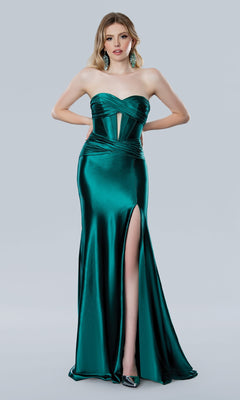 A woman models the Cut-Out Strapless Prom Gown: Stella Coture 25194 in emerald green satin, featuring a corset bodice, sheer panel accents, and a thigh-high slit, standing before a plain light gray backdrop.