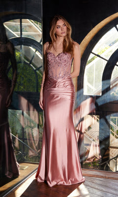 A woman stands by a window wearing the Juliet JT2531K sheer-bodice long prom dress, a mauve satin evening gown with a fitted embellished bodice and flowing skirt, styled for formal occasions.